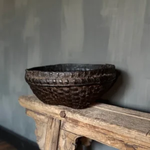 Oude leemmand met donker vlechtwerk en doorleefde uitstraling, geplaatst op een houten bankje in een landelijk interieur.