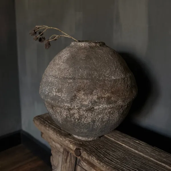 Handgemaakte papier-maché pot met doorleefde kalkachtige structuur op een houten kast in een sober wabi-sabi interieur.